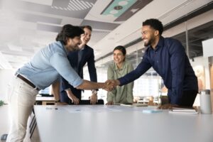 Cheerful positive multiethnic business colleagues celebrating corporate success, shaking hands over meeting table